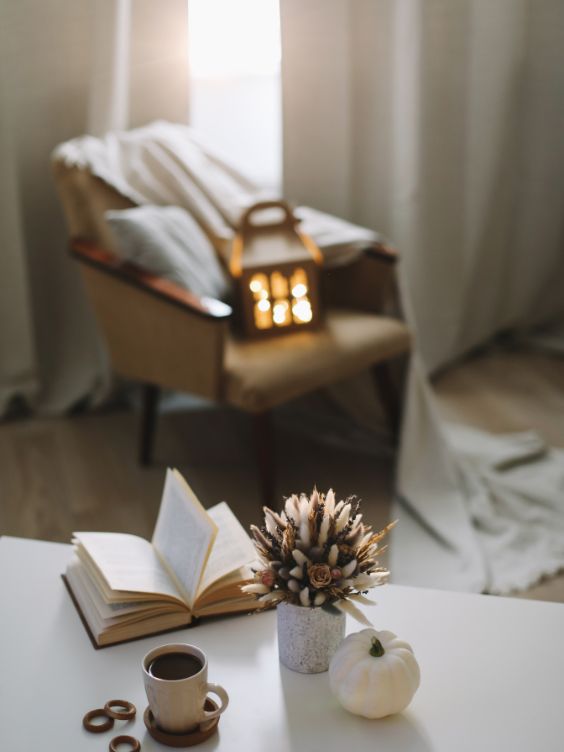 Coin lecture avec fauteuil, plaid, livre et tasse de café accompagnée d'u vase de fleurs et une mini citrouille blanche